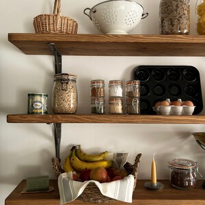 May include: Three wooden shelves with various kitchen items, including a colander, a basket of bananas and apples, a jar of coffee, and several jars of spices.