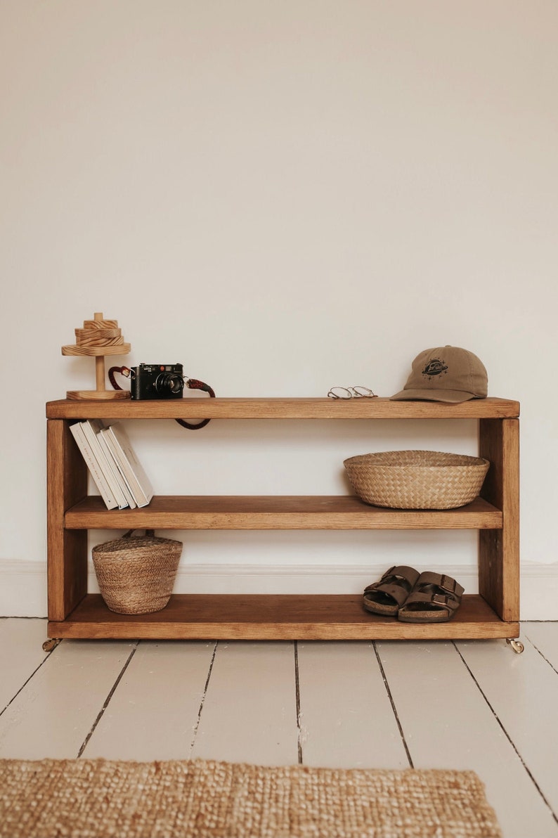 May include: A wooden shelf unit with three shelves, featuring a camera, a stack of books, two woven baskets, a pair of sandals, and a baseball cap. The shelf unit is on a white wooden floor.