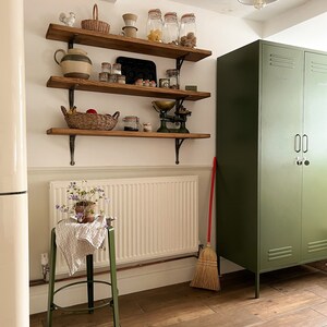 May include: A green metal cabinet with two doors and a white wall with wooden shelves filled with various kitchen items, including jars, a scale, and a basket. A broom leans against the cabinet.