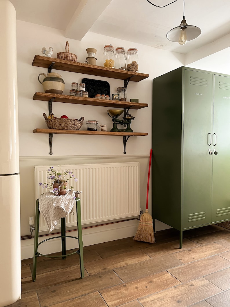 May include: A green metal cabinet with two doors and a white wall with wooden shelves filled with various kitchen items, including jars, a scale, and a basket. A broom leans against the cabinet.