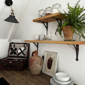 May include: A rustic kitchen with wooden shelves and black metal brackets. The shelves are decorated with glass jars, a potted fern, and a collection of glasses. A brown clay jug sits on the counter below the shelves, along with a wicker picnic basket and a framed print of a pitcher.