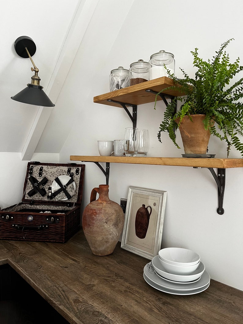 May include: A rustic kitchen with wooden shelves and black metal brackets. The shelves are decorated with glass jars, a potted fern, and a collection of glasses. A brown clay jug sits on the counter below the shelves, along with a wicker picnic basket and a framed print of a pitcher.