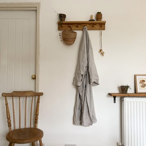 May include: A wooden coat rack with hooks and a shelf. A robe hangs on one of the hooks, and a small basket hangs on another. There are two small potted plants on the shelf.