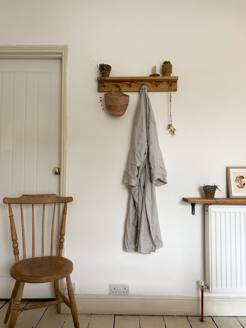 May include: A wooden coat rack with hooks and a shelf. A robe hangs on one of the hooks, and a small basket hangs on another. There are two small potted plants on the shelf.