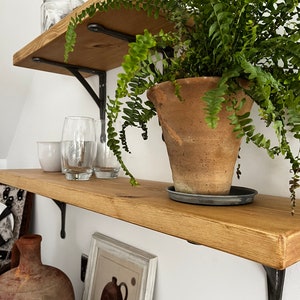May include: Two wooden shelves with black metal brackets, holding various items, including a potted fern, glass jars, and a glass. The shelves are mounted on a white wall.