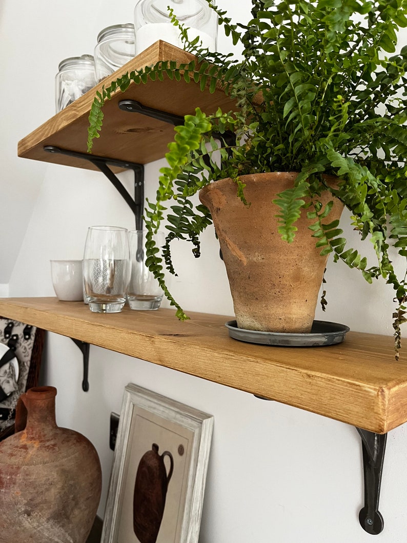 May include: Two wooden shelves with black metal brackets, holding various items, including a potted fern, glass jars, and a glass. The shelves are mounted on a white wall.