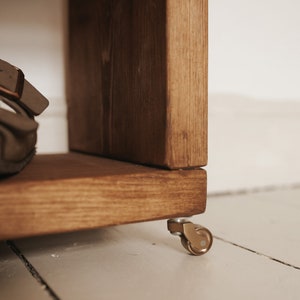 May include: A wooden shelf unit with a brown stain finish, featuring a single caster wheel on the bottom. The wheel is made of metal and has a gold finish.