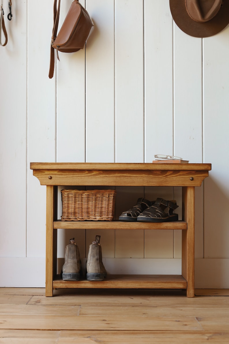 May include: A wooden shoe rack with two shelves. The top shelf has a wicker basket and a pair of sandals. The bottom shelf has two pairs of boots. The rack is against a white wall.