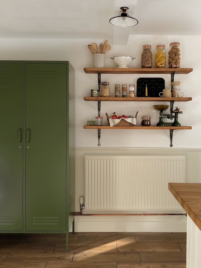 May include: A green metal locker cabinet with two doors and a set of three wooden shelves with various kitchen items, including jars of pasta, a white colander, and a vintage scale.