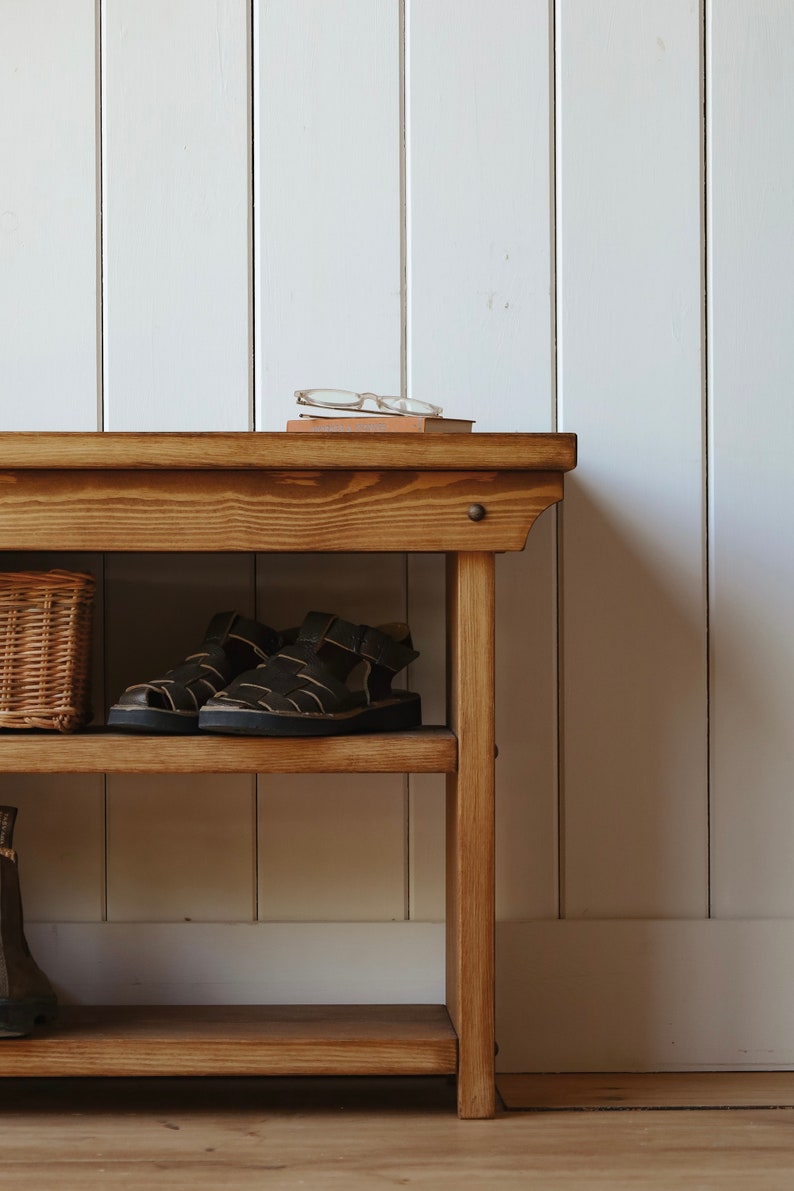 May include: A wooden shoe rack with two shelves. The top shelf has a pair of sandals and a book with glasses on top. The bottom shelf is empty. The shoe rack is in front of a white paneled wall.