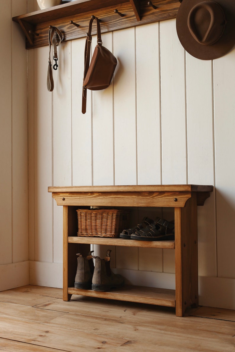 May include: A wooden shoe rack with two shelves. The top shelf has a wicker basket and a pair of sandals. The bottom shelf has two pairs of brown boots. The rack is against a white wall with a wooden shelf above it.