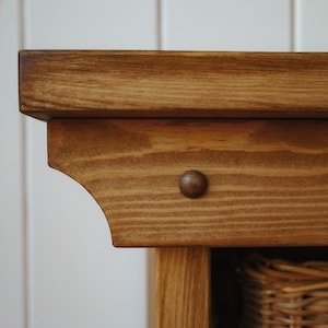 May include: Close-up of the top corner of a wooden cabinet with a brown stain. The cabinet has a decorative detail on the top edge and a small, round, brown knob.