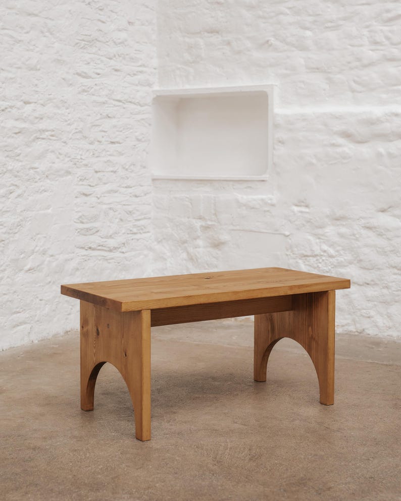 Pode incluir: Uma mesa de centro retangular de madeira com um acabamento natural em castanho claro. A mesa apresenta uma superf&iacute;cie superior plana e dois suportes laterais, cada um desenhado com um recorte arqueado distinto na base do m&oacute;vel.