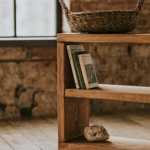 May include: A wooden shelf unit with two shelves, a woven basket on the top shelf, and a rock on the bottom shelf. The shelf unit is on wheels and has a rustic brown finish.