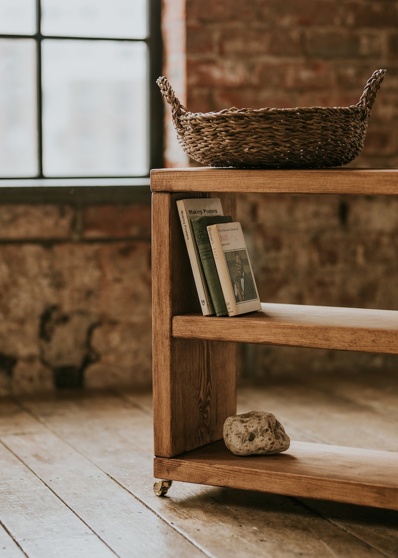 May include: A wooden shelf unit with two shelves, a woven basket on the top shelf, and a rock on the bottom shelf. The shelf unit is on wheels and has a rustic brown finish.