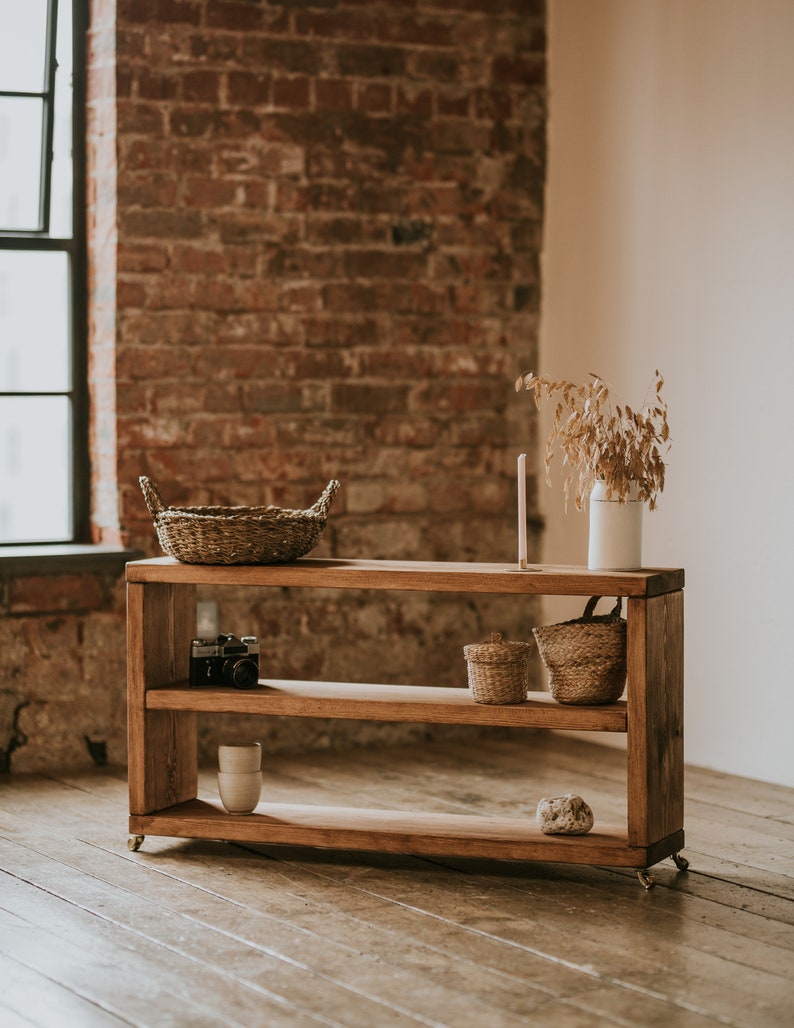 May include: A wooden shelf unit with three shelves, featuring a wicker basket, a camera, a ceramic mug, two wicker baskets, and a stone. The shelf unit is on a wooden floor in front of a brick wall.