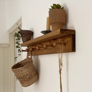 May include: A wooden shelf with hooks and a small shelf on top. There are two potted plants, a basket, and a bundle of dried grasses hanging from the hooks.