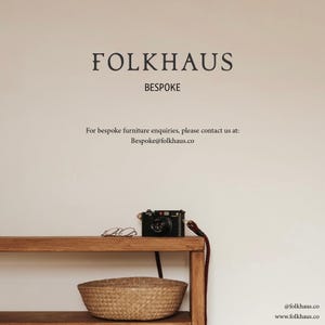 Pode incluir: Uma mesa de console de madeira com uma c&acirc;mera preta com al&ccedil;a estampada e um par de &oacute;culos na superf&iacute;cie superior, com uma cesta tran&ccedil;ada na prateleira inferior. A parede exibe o texto FOLKHAUS BESPOKE e informa&ccedil;&otilde;es de contato.