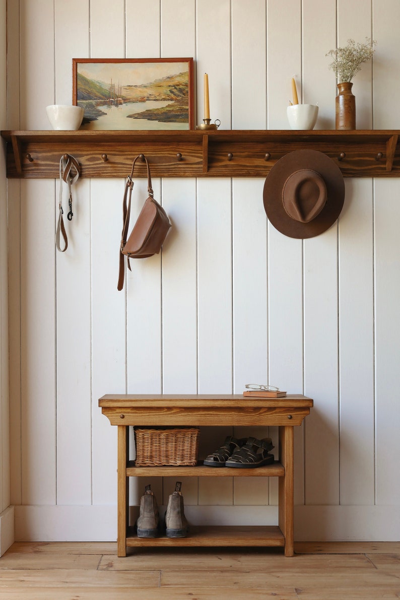 May include: A wooden entryway bench with two shelves, a woven basket, a pair of brown boots, and a pair of sandals. The bench is against a white paneled wall with a wooden shelf above it. The shelf has a small bowl, a framed picture, a hat, and two candlesticks.