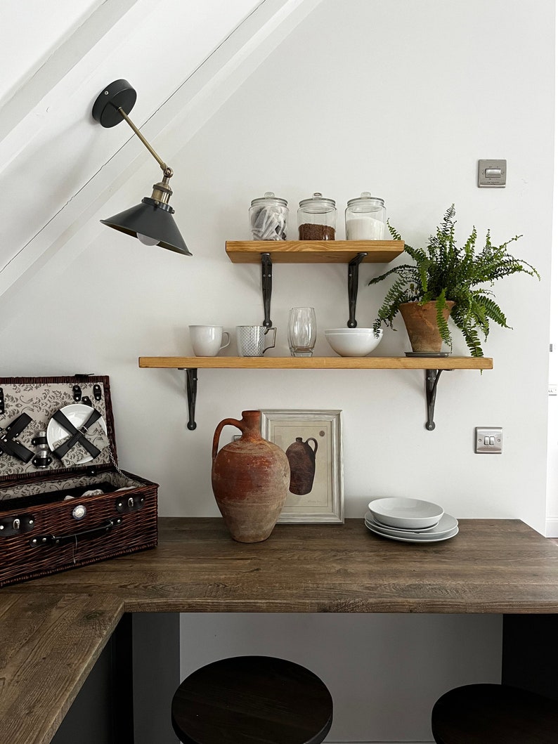 May include: A white wall with two wooden shelves with black metal brackets. The shelves are decorated with glass jars, a fern plant, a ceramic pitcher, a framed print, and white dishes. A brown wooden countertop with a black corner and two black stools is in the foreground.