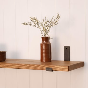 Op de afbeelding: Een rustiek houten plank met een bruine keramische vaas met gedroogde bloemen. De plank wordt ondersteund door donkere metalen beugels. De achtergrond is een witte wand met panelen. Minimalistisch, natuurlijk interieur.