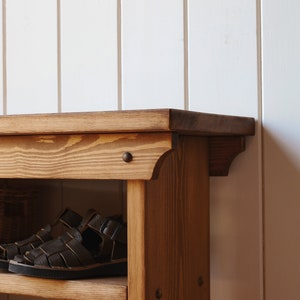 May include: A wooden shoe rack with two shelves and a top surface. The rack is made of natural wood with a brown stain. There are two pairs of brown leather sandals on the top shelf.