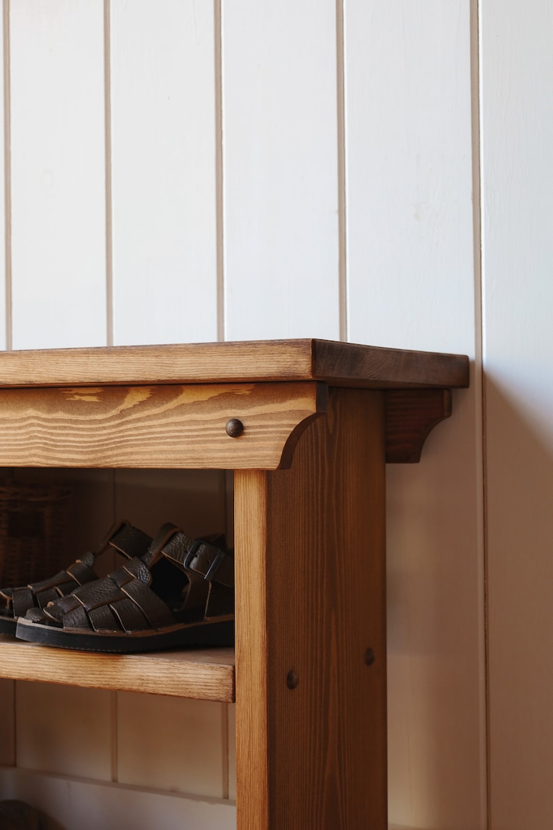 May include: A wooden shoe rack with two shelves and a top surface. The rack is made of natural wood with a brown stain. There are two pairs of brown leather sandals on the top shelf.
