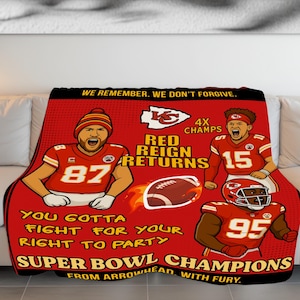 May include: Red and black blanket featuring cartoon illustrations of football players. The blanket has the text "WE REMEMBER, WE DON'T FORGIVE" and "SUPER BOWL CHAMPIONS". The blanket is displayed on a white couch.