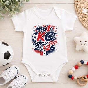 Op de afbeelding: Een wit babyrompertje met korte mouwen en een grafische print met de tekst MY 1ST KC WORLD CUP 2026 in rode en marineblauwe letters. Het ontwerp bevat een voetbal, een wereldbol-icoon en sterren, met drie drukknopen aan de onderkant.