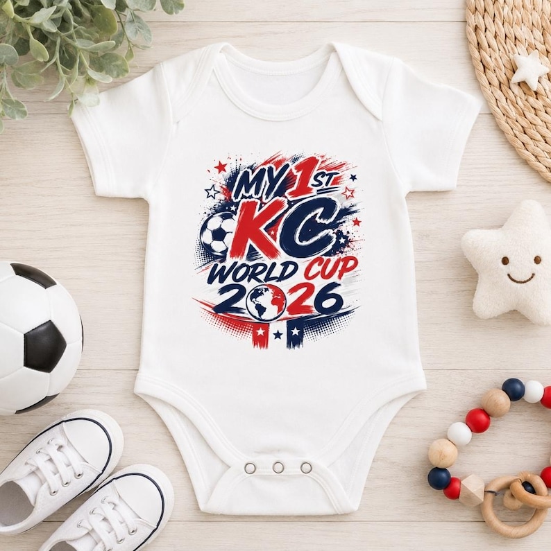 Op de afbeelding: Een wit babyrompertje met korte mouwen en een grafische print met de tekst MY 1ST KC WORLD CUP 2026 in rode en marineblauwe letters. Het ontwerp bevat een voetbal, een wereldbol-icoon en sterren, met drie drukknopen aan de onderkant.