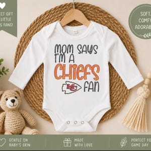 May include: White baby bodysuit with long sleeves, featuring the text "MOM SAYS I'M A CHIEFS FAN" in black and orange. The Kansas City Chiefs logo is also displayed. The bodysuit is hanging on a wooden hanger.