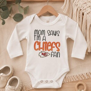 May include: White baby bodysuit with long sleeves, featuring the text "MOM SAYS I'M A CHIEFS FAN" in black, orange, and red. The design includes the Kansas City Chiefs logo. The bodysuit is hanging on a wooden hanger.