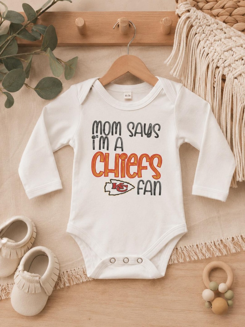 May include: White baby bodysuit with long sleeves, featuring the text "MOM SAYS I'M A CHIEFS FAN" in black, orange, and red. The design includes the Kansas City Chiefs logo. The bodysuit is hanging on a wooden hanger.