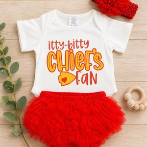 May include: A white baby bodysuit with the text "itty-bitty CHIEF'S FAN" in red and yellow. The outfit includes a red tutu diaper cover and a red floral headband. A wooden teething ring is also present.