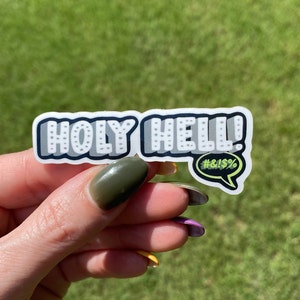 Puede incluir: Una pegatina blanca con las palabras "HOLY HELL!" en una fuente audaz y estilizada. La pegatina tiene un contorno negro y un bocadillo con los símbolos "#&!$" en verde.
