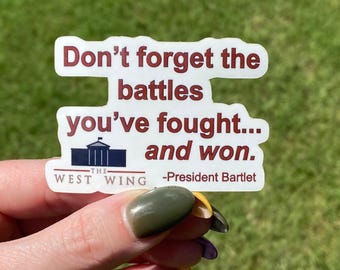 May include: A white die-cut sticker featuring the dark red text Don't forget the battles you've fought... and won. Below the text is a dark blue icon of the White House, the words THE WEST WING, and the attribution -President Bartlet in red.