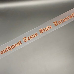 May include: A clear plastic ribbon with the words "Southwest Texas State University" printed in brown lettering. The ribbon has a gold star seal on each end.