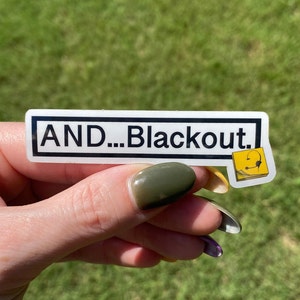 Puede incluir: Una pegatina blanca con texto negro que dice "AND...Blackout." Una peque&ntilde;a pegatina amarilla con un icono de auriculares negros est&aacute; a la derecha de la pegatina m&aacute;s grande.