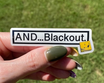 May include: A white sticker with black text that reads "AND...Blackout." A small yellow sticker with a black headset icon is to the right of the larger sticker.