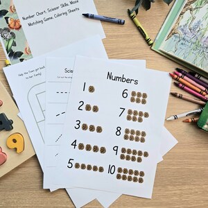 May include: A set of printable worksheets for kids to learn numbers. The worksheets feature a number and a corresponding number of brown circles. The worksheets are on a wooden table with a box of crayons and a wooden number puzzle.
