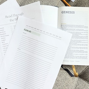 May include: A white sheet of paper with the title "Read Through the Bible" and a checklist of books of the Bible. A calendar with the months of the year is printed on the sheet. The word "Genesis" is written in green. A Bible with the title "Genesis" is open to the first chapter.