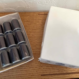 May include: A package of 8 spools of black thread in a clear plastic container. The spools are arranged in two rows of four. The package is sitting on a wooden surface.