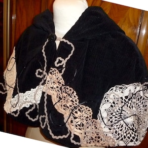 May include: Black corduroy hooded cape with white and beige lace trim. The lace is sewn in a decorative pattern.