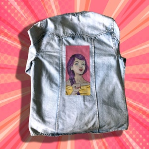 May include: A light blue denim jacket with a painted image of a woman with purple hair and a yellow shirt on the back.
