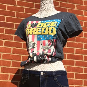 K&ouml;nnte beinhalten: Ein dunkelgraues T-Shirt mit einem Comic-Grafik-Design, das den Text "JUDGE DREDD" in Gelb und Blau vor einem amerikanischen Flaggen-Design zeigt. Das Shirt wird mit dunkelblauen Jeansshorts kombiniert.