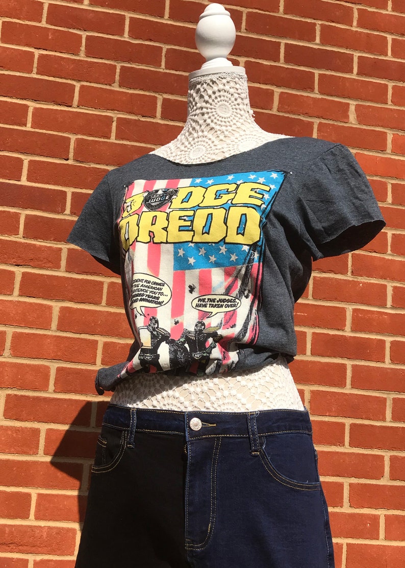 K&ouml;nnte beinhalten: Ein dunkelgraues T-Shirt mit einem Comic-Grafik-Design, das den Text "JUDGE DREDD" in Gelb und Blau vor einem amerikanischen Flaggen-Design zeigt. Das Shirt wird mit dunkelblauen Jeansshorts kombiniert.