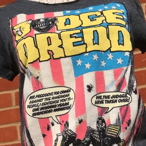 K&ouml;nnte beinhalten: Dunkelgraues T-Shirt mit einem Judge Dredd-Motiv, das den Text "JUDGE DREDD" in Gelb und Blau zeigt. Das Design beinhaltet eine amerikanische Flagge und Comic-Sprechblasen. Das T-Shirt hat kurze &Auml;rmel.