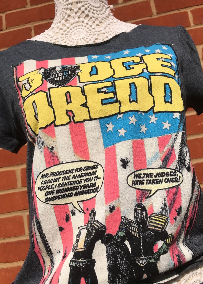 K&ouml;nnte beinhalten: Dunkelgraues T-Shirt mit einem Judge Dredd-Motiv, das den Text "JUDGE DREDD" in Gelb und Blau zeigt. Das Design beinhaltet eine amerikanische Flagge und Comic-Sprechblasen. Das T-Shirt hat kurze &Auml;rmel.
