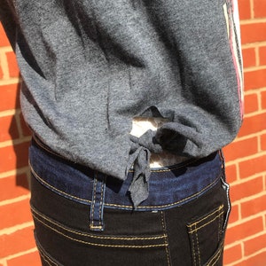 K&ouml;nnte beinhalten: Ein graues Oberteil mit Bindeband, kombiniert mit dunkel gewaschenen Jeans. Die Jeans hat gelbe N&auml;hte und einen dunkelblauen Bund. Das Oberteil hat einen rosa-wei&szlig;en Streifen an der Seite.