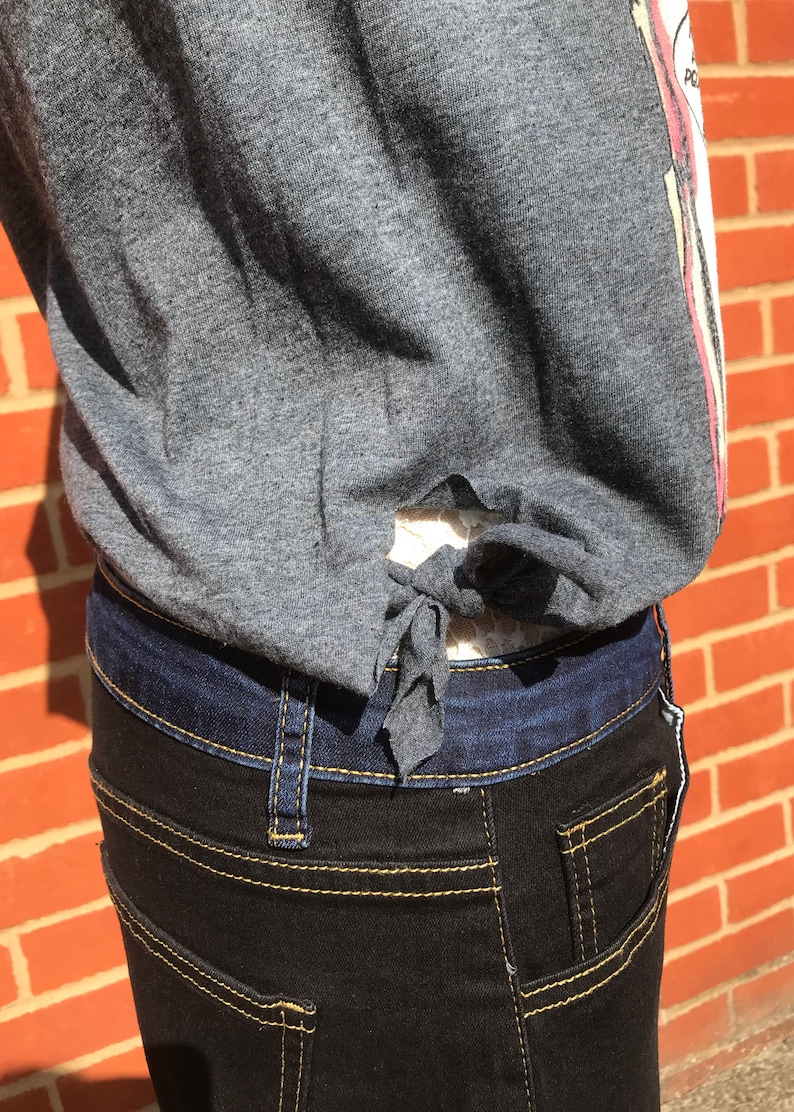 K&ouml;nnte beinhalten: Ein graues Oberteil mit Bindeband, kombiniert mit dunkel gewaschenen Jeans. Die Jeans hat gelbe N&auml;hte und einen dunkelblauen Bund. Das Oberteil hat einen rosa-wei&szlig;en Streifen an der Seite.